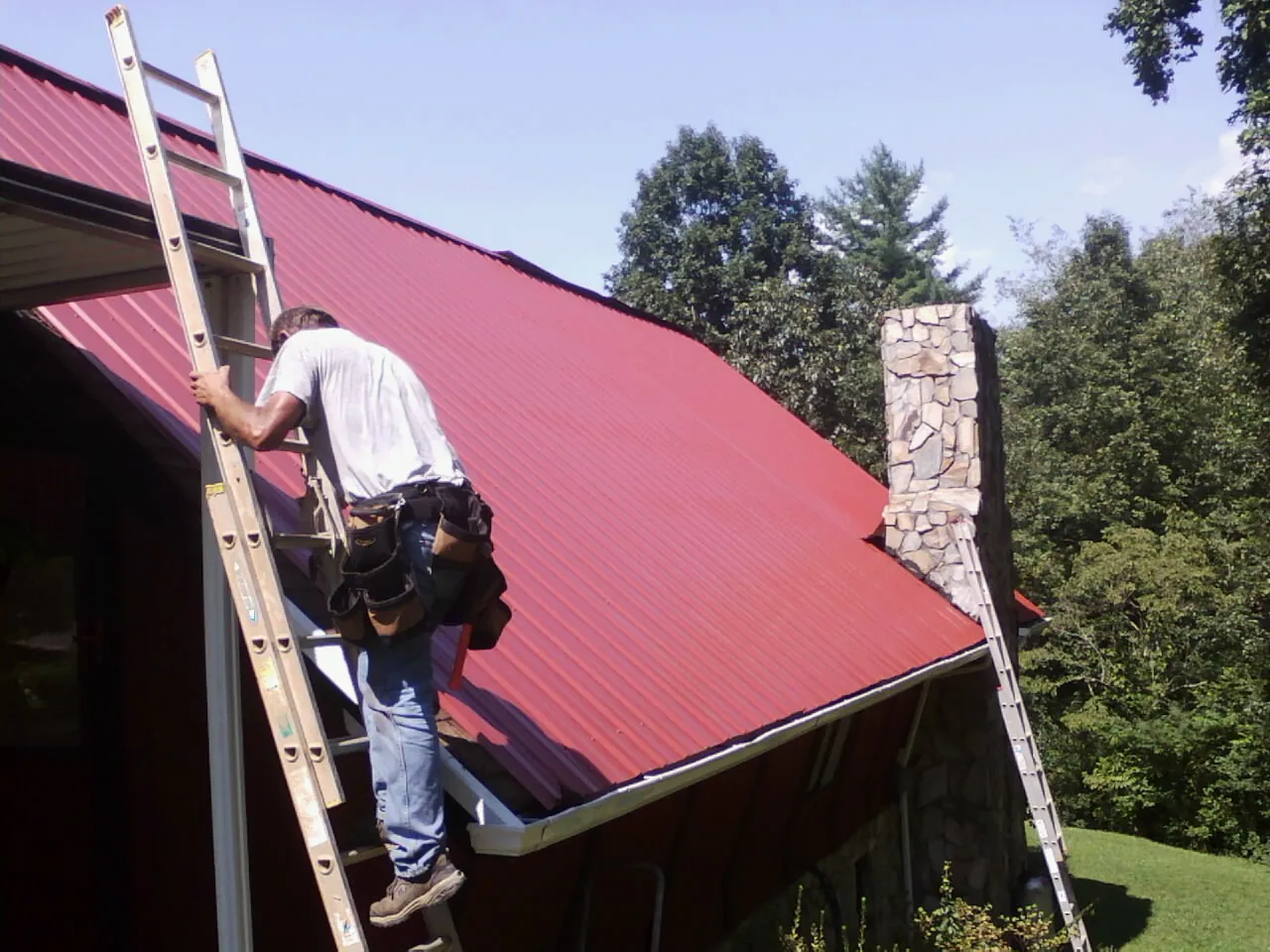 Roofer on a ladder beside a red metal roof and stone chimney — Soffit & Fascia Repair by Roofing Brothers in Dearing